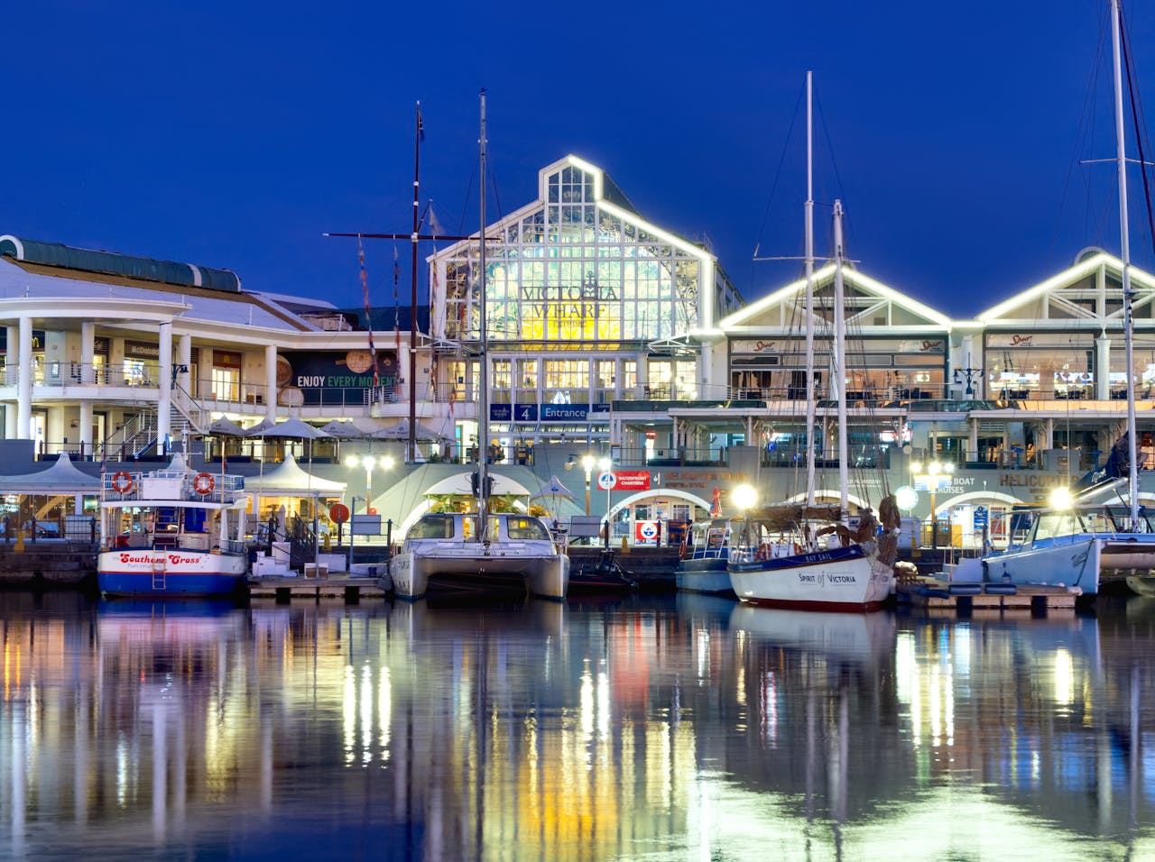 services-04 Illuminated view of Victoria Wharf Shopping Centre surrounded by yachts and boats at night in Cape Town, South Africa.